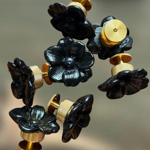 Puede incluir: Un conjunto de cinco pomos negros con forma de flor, cada uno con un diseño de pétalos tallados y brillantes, y una base cilíndrica de metal dorado con un borde texturizado y moleteado para un mejor agarre, dispuestos en un grupo.