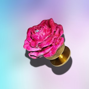 May include: A decorative cabinet knob featuring a sculpted, vibrant magenta rose with a single small, clear crystal embedded in the center. The rose is attached to a circular, gold-toned metal base with a textured, knurled edge for grip.