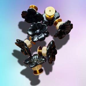 May include: A collection of five drawer knobs, each featuring a glossy black flower-shaped front with detailed petals. The base of each knob is made of metallic gold-toned hardware with a textured, knurled cylindrical grip for easy handling.