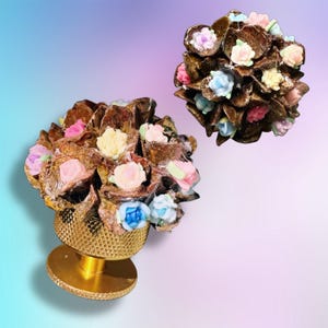 May include: A decorative knob featuring a gold-toned metal base with a textured, diamond-patterned surface. The top is adorned with a cluster of dark brown, shell-like elements interspersed with small, multi-colored pastel floral accents.