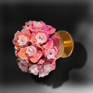 May include: A decorative cabinet knob featuring a cluster of textured pink and orange floral shapes with small, clear, faceted crystal centers. The knob is attached to a circular, gold-toned metal base with a knurled edge for grip and installation.