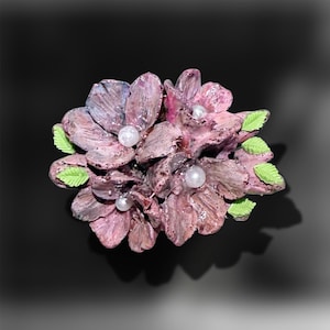Può includere: Una spilla floreale con molteplici petali rosa polvere testurizzati e finitura lucida. Tre piccole perle bianche rotonde e artificiali sono incastonate al centro del gruppo di fiori. Diverse piccole foglie verde brillante con bordi seghettati sono intervallate tra i petali.