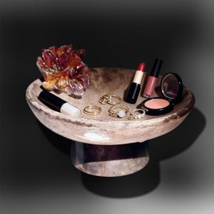 Könnte beinhalten: Eine Fußschale aus gesprenkeltem braunem Stein, die ein Bündel aus violetten und orangefarbenen Kristallen, drei goldfarbene Ringe, einen schwarzen Lippenstift, einen rosa Lipgloss und eine runde Puderdose mit schwarzem Deckel enthält.