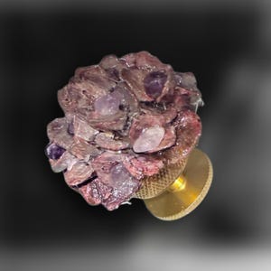 Può includere: Un pomello decorativo per mobili con una parte superiore a forma di fiore composta da petali in resina traslucida viola e rosa, incastonati con piccoli cristalli di ametista e quarzo chiaro, fissato su una base cilindrica in ottone testurizzato.