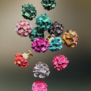 May include: A collection of spherical floral-shaped knobs featuring textured petals in various colors including green, pink, red, orange, brown, and teal. Each knob has a metallic gold-toned cylindrical base with a knurled edge for attachment.