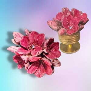 May include: Two decorative knobs featuring clusters of deep pink, multi-petaled flowers. Each flower cluster is accented with a small, clear, central rhinestone. The knobs are mounted on cylindrical, gold-toned metal bases with a textured grip.