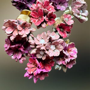 Könnte beinhalten: Eine Sammlung dekorativer Schubladenknöpfe aus handbemalten Tannenzapfensegmenten, die wie blühende Blumen in Magenta, Dunkelviolett und zartem Rosa geformt sind und jeweils auf einem zylindrischen Sockel aus Gold- oder Silbermetall sitzen.