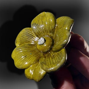 May include: A decorative drawer knob shaped like a five-petaled flower with a glossy, mustard-yellow glazed finish. The petals feature subtle vertical ridges and a dark, textured center. The knob has a metallic base and a reflective surface.