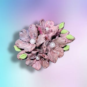 May include: A decorative brooch featuring a cluster of textured, dusty pink flowers with visible brushstroke details. Each flower center is accented with a small, round, white pearl bead. Several small, bright lime green leaves are tucked between the petals.