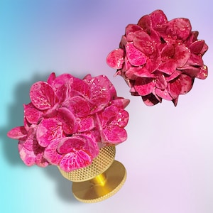 May include: Two decorative knobs featuring vibrant, glossy magenta hydrangea flower heads attached to circular, textured gold-toned metal bases. The petals exhibit a realistic, layered structure with a shiny finish, contrasting with the metallic hardware.