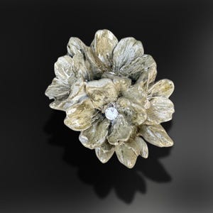 May include: A ring featuring a sculpted, multi-layered flower design in a muted, earthy metallic tone. The center of the floral arrangement is accented with a single, small, clear, round-cut gemstone that catches the light.