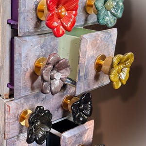 May include: A small storage unit with multiple drawers featuring a marbled gray finish. Each drawer is equipped with a gold-toned metal knob shaped like a flower, available in various colors including red, green, yellow, and black.