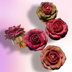 Könnte beinhalten: Eine Auswahl an rosenförmigen Schrankknöpfen in verschiedenen Farben, darunter tiefrot, burgunderrot und braun. Ein Knopf ist an einer goldfarbenen Metallbasis befestigt. Jede Rose hat ein detailliertes, strukturiertes Aussehen, bei einigen mit einem kleinen Edelstein in der Mitte.