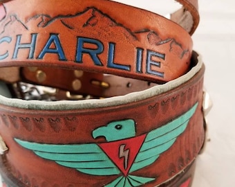 May include: Stacked brown leather belts with hand-painted designs. One belt features the name "CHARLIE" in blue, another has a turquoise eagle with a red lightning bolt, and another has a mountain range. The belts have silver rivets and colorful accents.