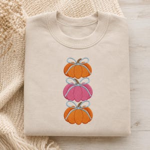 Op de afbeelding: Een beige sweater met ronde hals voorzien van een geborduurd ontwerp van drie gestapelde pompoenen. De bovenste en onderste pompoen zijn oranje, terwijl de middelste pompoen roze is. Elke pompoen is gestrikt met een lichtblauw lint.
