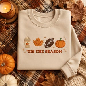 May include: A beige crewneck sweatshirt featuring embroidered autumn-themed icons including a coffee cup, a maple leaf, a football, and a pumpkin. The text 'TIS THE SEASON is embroidered in orange below the icons, accented by small star shapes.