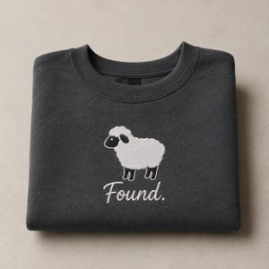 May include: A charcoal gray crewneck sweatshirt featuring an embroidered white sheep graphic centered on the chest. Below the sheep, the word Found is embroidered in a white cursive script font. The garment has a ribbed collar and long sleeves.