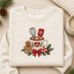 Puede incluir: Una sudadera de cuello redondo color crema con un bordado de una taza blanca que contiene un batidor y una espátula roja. La taza está decorada con un corazón rojo y un copo de nieve. Debajo hay un hombre de jengibre, un lazo rojo y acebo.
