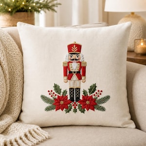 Può includere: Un cuscino quadrato color bianco sporco con un ricamo centrale raffigurante uno schiaccianoci in uniforme rossa con dettagli dorati, affiancato da fiori di stella di Natale rossi, aghi di pino verdi e piccole bacche rosse alla base.