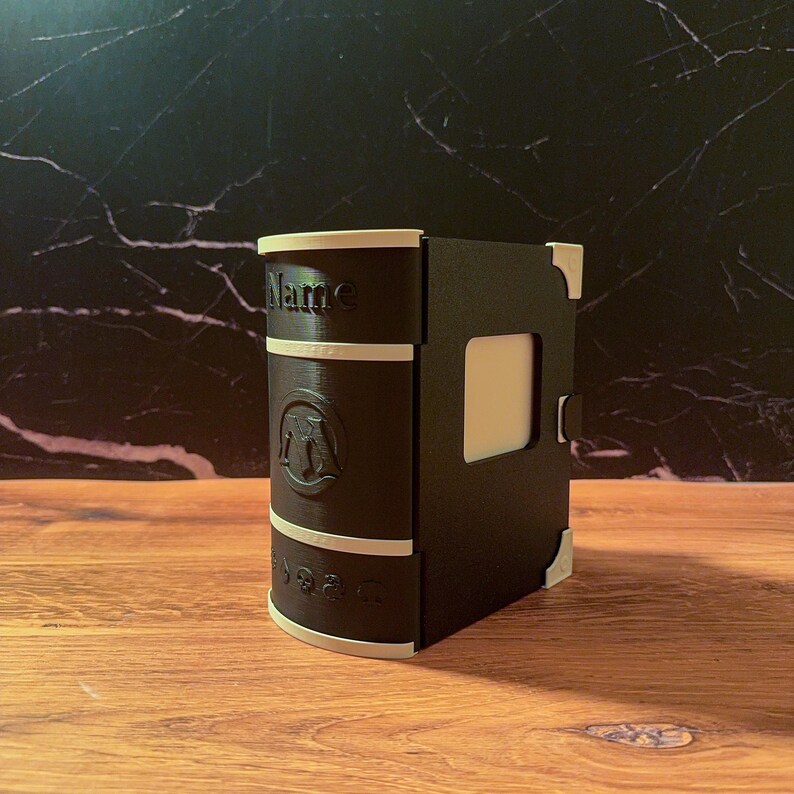 May include: A black and off-white 3D printed deck box shaped like a book. The spine features the word Name, a circular emblem with the letter M, and a row of five small icons. The box has light-colored corner accents and a rectangular side panel.