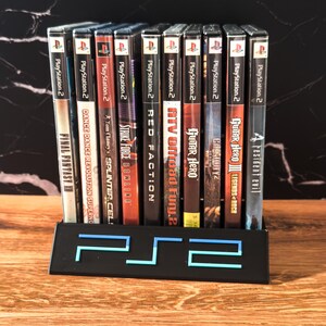 May include: A selection of PlayStation 2 game cases in a black stand with a blue illuminated "PS2" logo. Titles include "Final Fantasy XI", "Guitar Hero", and "Resident Evil". The games are in their original packaging.