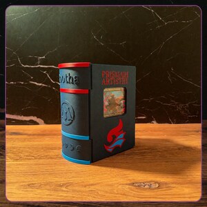 K&ouml;nnte beinhalten: Eine dunkelgraue, rechteckige Kartenaufbewahrungsbox in Buchform mit einem roten und blauen Buchr&uuml;cken. Die Vorderseite zeigt den roten Schriftzug PRISMARI ARTISTRY, ein rot-blaues Flammenemblem und ein kleines quadratisches Sichtfenster mit einer Karte.