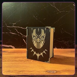May include: A black rectangular box with a hinged side, featuring a stylized gold and black mask graphic on the front cover. The box has metallic corner accents and a light-colored interior visible on the side, presenting a sleek, structured design.