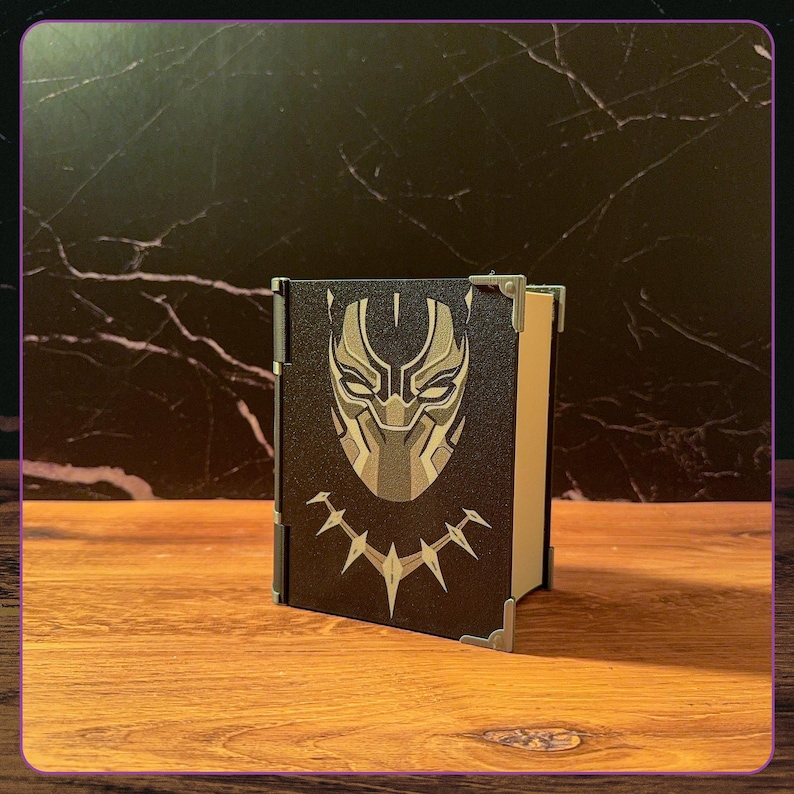 May include: A black rectangular box with a hinged side, featuring a stylized gold and black mask graphic on the front cover. The box has metallic corner accents and a light-colored interior visible on the side, presenting a sleek, structured design.
