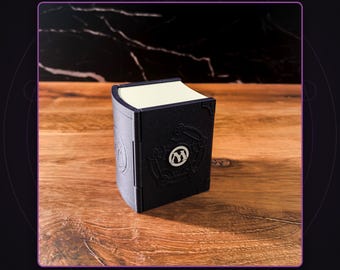 May include: A black rectangular storage box designed to resemble a book with a curved spine. The front cover features a circular emblem with the letter M in the center, surrounded by embossed geometric lines. The top edge shows cream-colored pages.