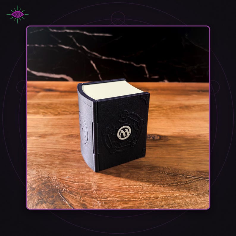 May include: A black rectangular storage box designed to resemble a book with a curved spine. The front cover features a circular emblem with the letter M in the center, surrounded by embossed geometric lines. The top edge shows cream-colored pages.