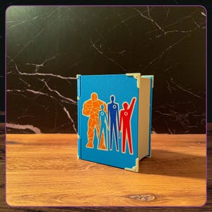 May include: A blue rectangular storage box featuring a graphic of four stylized figures in orange, blue, and red. The box has cream-colored corner accents and a hinge on the left side. The figures are arranged in a row, each with a circular emblem.