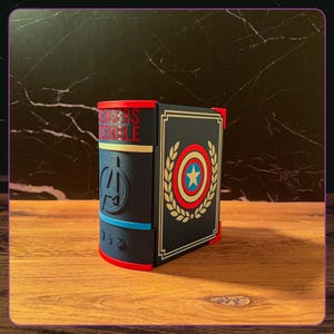 May include: A decorative book-shaped storage box featuring a dark blue spine with the red text AVENGERS ASSEMBLE, an Avengers logo, and small icons. The front cover is dark blue with a gold laurel wreath surrounding a red, white, and blue shield.