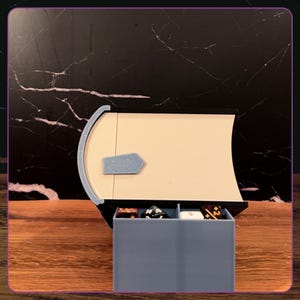 May include: A book-shaped storage box with a cream-colored spine and a light blue accent piece. The box features a pull-out drawer at the bottom, divided into two compartments, containing several multi-sided gaming dice in various colors.