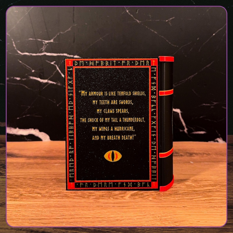May include: A black rectangular book-shaped object with red trim and a spine featuring three red bands. The cover displays the text: "My armour is like tenfold shields, my teeth are swords, my claws spears, the shock of my tail a thunderbolt, my wings a hurricane, and my breath death!" with a yellow and red eye icon below.