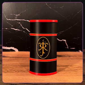 May include: A cylindrical black container featuring a red rim at the top and bottom, with two additional red horizontal bands encircling the middle. A gold oval emblem is centered on the front, displaying the stylized JR Tolkien monogram logo.