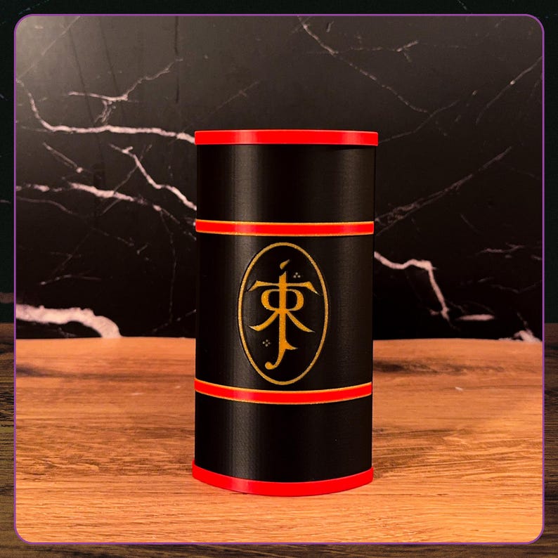 May include: A cylindrical black container featuring a red rim at the top and bottom, with two additional red horizontal bands encircling the middle. A gold oval emblem is centered on the front, displaying the stylized JR Tolkien monogram logo.