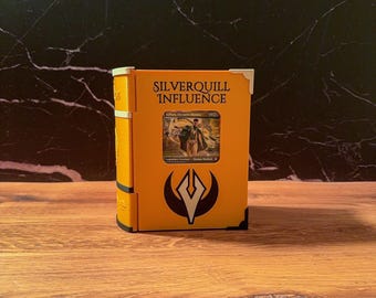 May include: A bright yellow, book-shaped storage case featuring the text Silverquill Influence at the top and a black emblem below. A rectangular cutout in the center displays a trading card depicting a figure in a suit. The case has white corner accents.