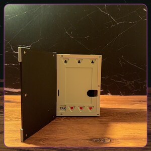 May include: A rectangular white plastic storage box with a hinged black lid. The front panel features the word TAX printed in black, three small red heart icons, and three keyhole-shaped mounting slots along the top edge of the interior frame.