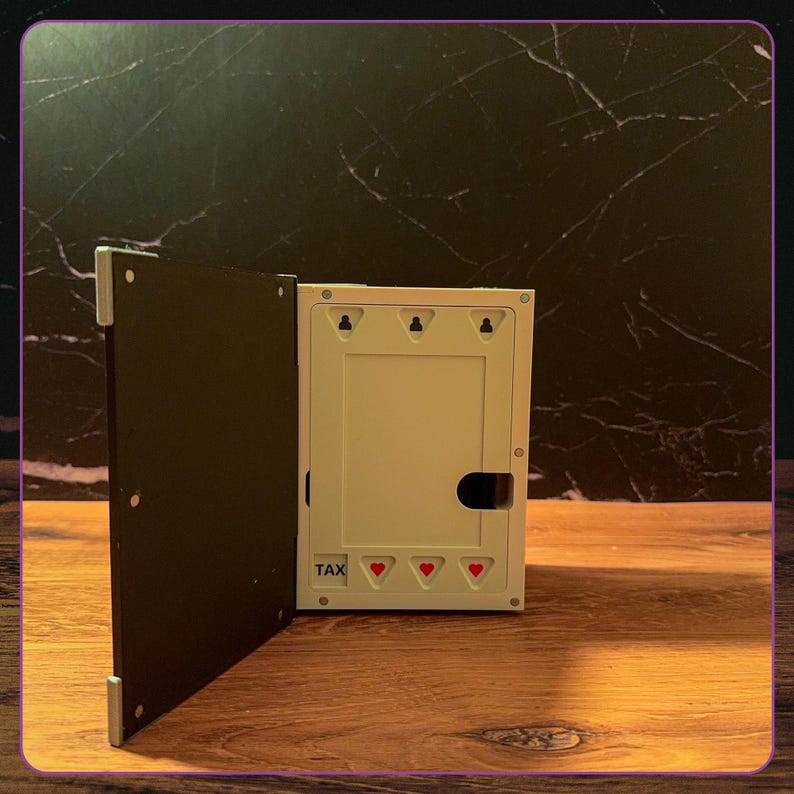 May include: A rectangular white plastic storage box with a hinged black lid. The front panel features the word TAX printed in black, three small red heart icons, and three keyhole-shaped mounting slots along the top edge of the interior frame.