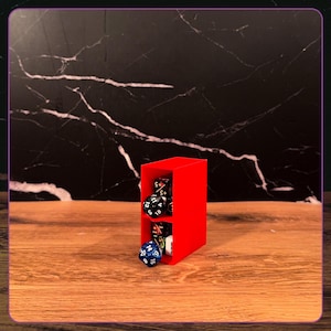 May include: A bright red, rectangular, two-tiered storage tower made of plastic, designed to hold polyhedral dice. The tower features an open front, with several multi-sided gaming dice in black, blue, and white colors resting inside the compartments.