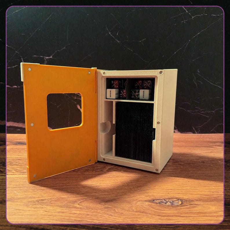May include: A rectangular white storage box with an open orange door featuring a square cutout. The interior contains a vertical compartment filled with a deck of black cards and a top shelf holding four small dice with white and dark markings.