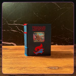 K&ouml;nnte beinhalten: Eine schwarze Deck-Box mit dem roten Schriftzug PRISMARI ARTISTRY, einem zentralen Sichtfenster f&uuml;r eine Kreaturenkarte und einem rot-blauen flammenartigen Emblem darunter. Die Seite der Box ist blau mit roten Akzenten.