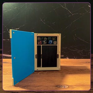 May include: A rectangular storage box with a bright blue hinged door and a cream-colored frame. The interior features a top compartment filled with various multi-sided gaming dice and a larger lower section containing a deck of black playing cards.