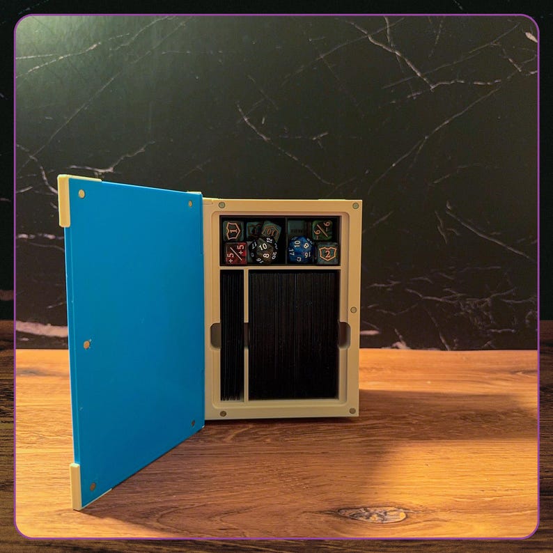 May include: A rectangular storage box with a bright blue hinged door and a cream-colored frame. The interior features a top compartment filled with various multi-sided gaming dice and a larger lower section containing a deck of black playing cards.