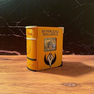 May include: A yellow 3D-printed deck box shaped like a book with the text Silverquill Influence printed on the front. The box features a central window displaying a trading card, a black and white emblem, and decorative corner accents.