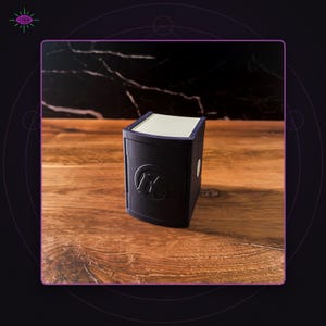 May include: A matte black rectangular storage box with a curved front panel featuring a circular embossed logo with the letter M. The box has a light-colored top lid and a smooth, solid construction, designed as a compact container for small items.