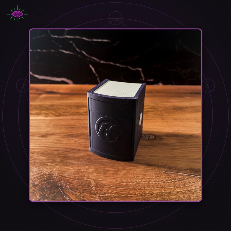 May include: A matte black rectangular storage box with a curved front panel featuring a circular embossed logo with the letter M. The box has a light-colored top lid and a smooth, solid construction, designed as a compact container for small items.