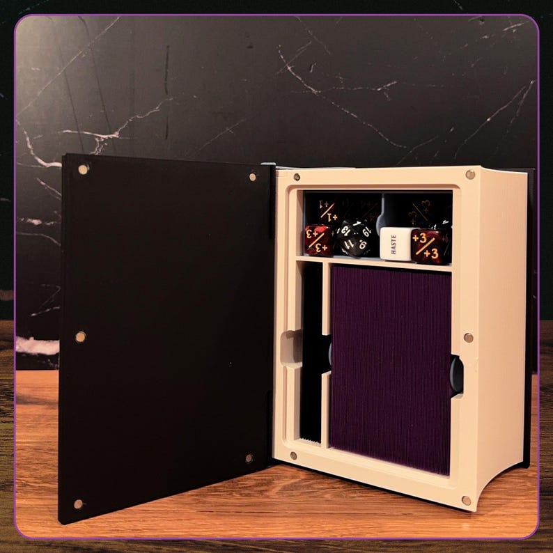 May include: A rectangular storage box with a black hinged cover and a cream-colored interior frame. The box contains a deck of purple-edged cards and several multi-sided gaming dice, including one white die with the word HASTE printed on it.