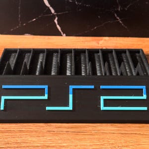 May include: Black PS2 game holder with blue and light blue detailing. The holder features multiple slots for game storage. The letters "PS2" are displayed on the front. The background is a wooden surface with a black marble pattern.