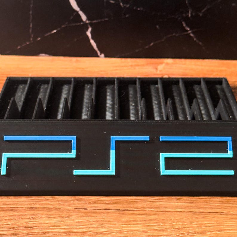 May include: Black PS2 game holder with blue and light blue detailing. The holder features multiple slots for game storage. The letters "PS2" are displayed on the front. The background is a wooden surface with a black marble pattern.